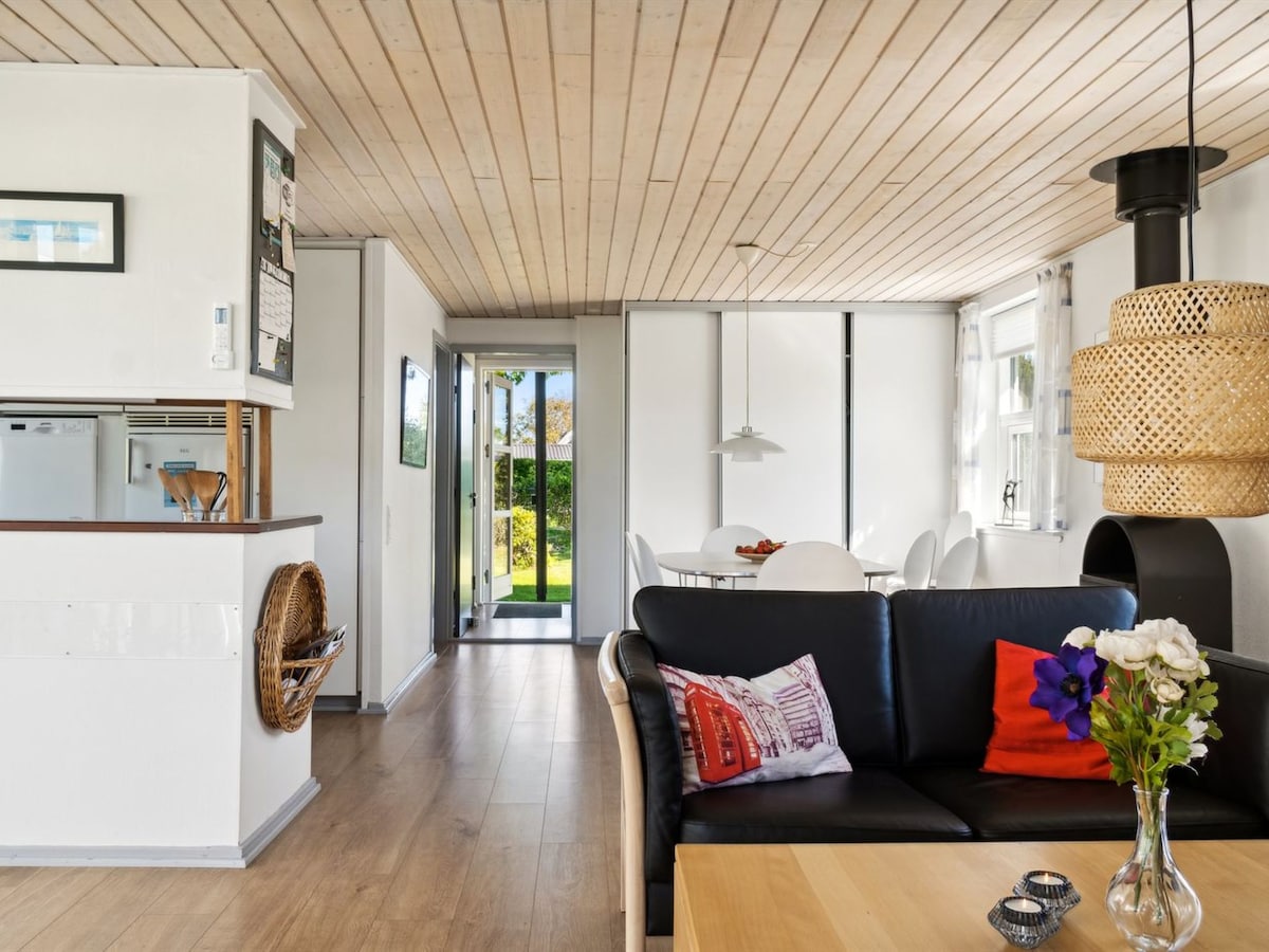 An open living space features a comfortable black sofa complemented by a wooden coffee table. A bright vase holds fresh flowers, and large windows allow natural light to fill the room. A dining area with white chairs and a light fixture is visible in the background.