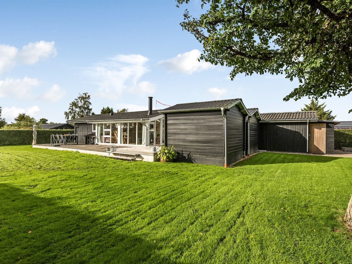 The exterior of the house is presented, showcasing a modern design with a large lawn that is neatly maintained. A patio area with outdoor seating is visible, along with large windows that offer views of the surrounding greenery and provide ample natural light.