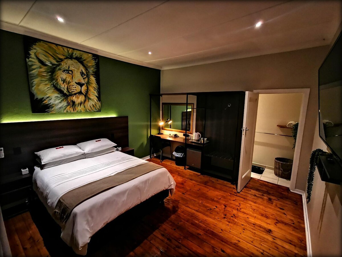 The room features a spacious bed with light-colored linens, accompanied by a large artwork of a lion on the green accent wall. A desk space with a mirror is visible, along with a wooden floor that adds warmth to the area. A television is mounted on the opposite wall.