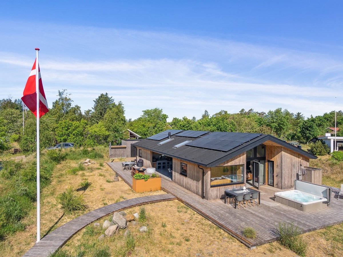 A single-family house features a wooden exterior with a modern design. Solar panels are installed on the sloped roof, while a wooden terrace surrounds the building. A hot tub is positioned on the terrace, set among natural landscaping, with a flagpole displaying a flag nearby.