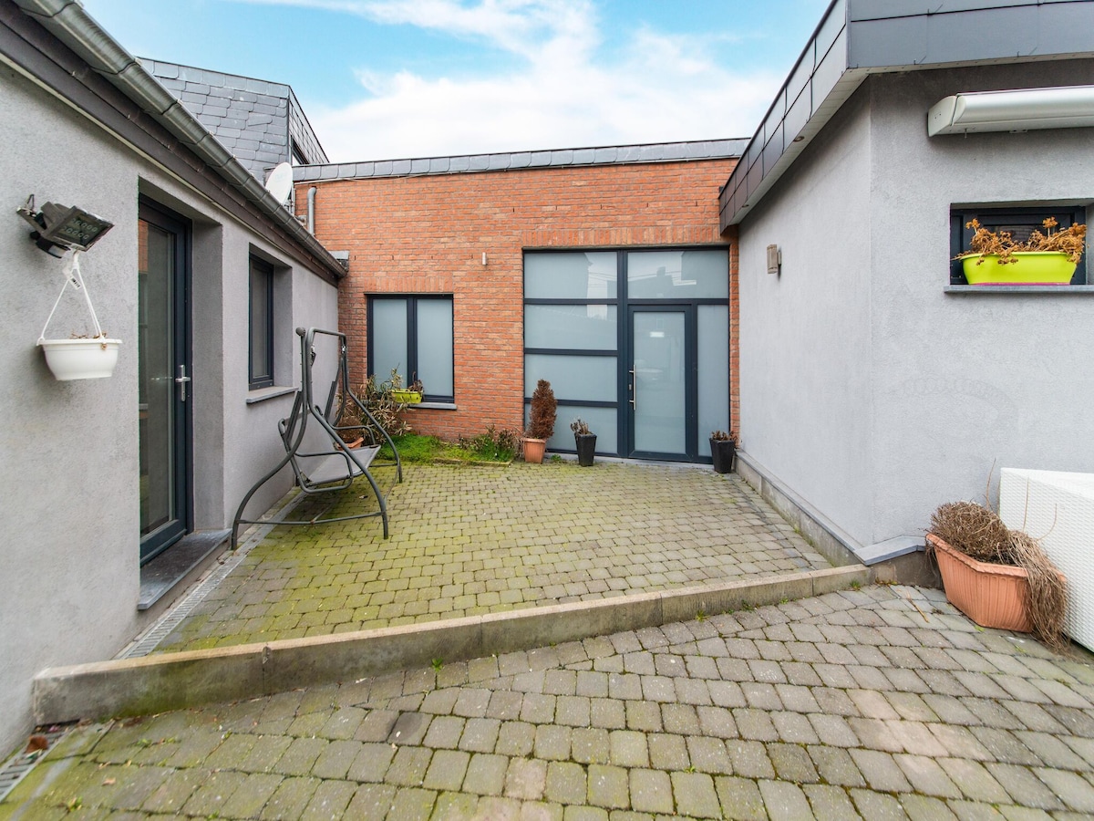 An enclosed courtyard features a combination of paved stones and greenery. A small decorative plant is positioned near the entrance, while a hanging planter can be seen on the adjacent wall. Natural light illuminates the area, highlighting the modern architecture.