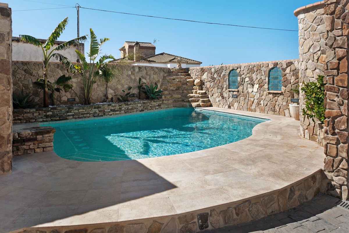 A spacious outdoor pool is surrounded by natural stone walls, featuring steps that lead into the water. Lush tropical plants are positioned along the edges, and sunlight reflects off the clear blue surface, creating an inviting spot for relaxation.
