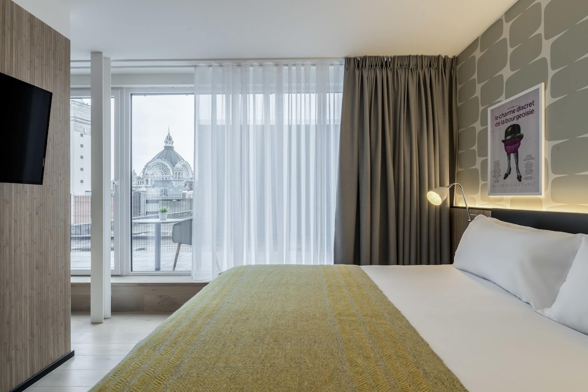 A well-lit hotel room features a spacious bed with crisp linens and a textured yellow throw. Floor-to-ceiling windows provide natural light and showcase a view of nearby architectural details, with sheer curtains gently framing the scene.