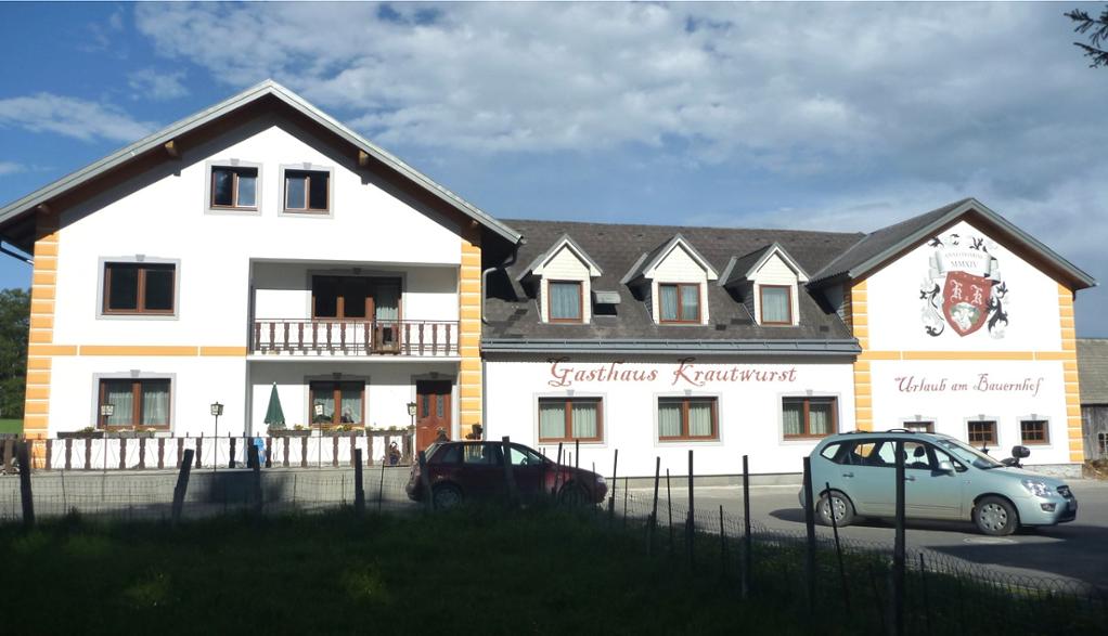 The exterior of a cozy guesthouse is presented, featuring a two-story building with a tiled roof and welcoming balconies. The white walls are accented by orange detailing, and the establishment's name is visible across the front. Cars are parked nearby, and a grassy area is in the foreground.