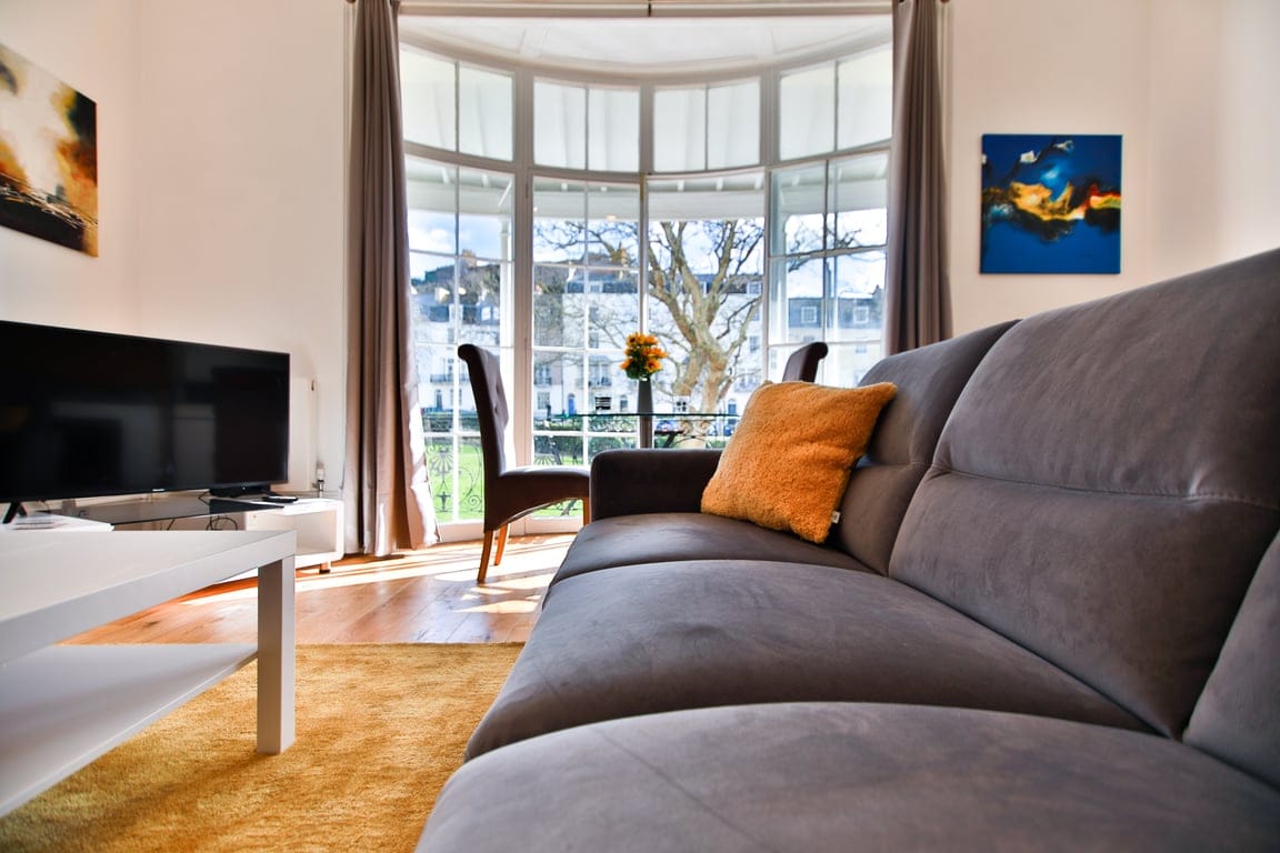 A spacious living area is accentuated by large bay windows that allow natural light to fill the space. A comfortable gray sofa is positioned invitingly, complemented by a vibrant accent pillow. A sleek white coffee table adds a modern touch, with a flat-screen TV nearby.