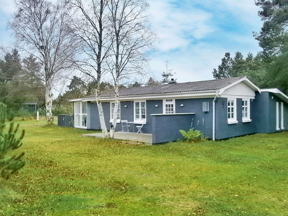 The single-family house, surrounded by natural greenery, features a blue exterior with large windows. A small terrace is visible in the front, complemented by two seated chairs. Mature trees and open lawn space create a peaceful outdoor environment.