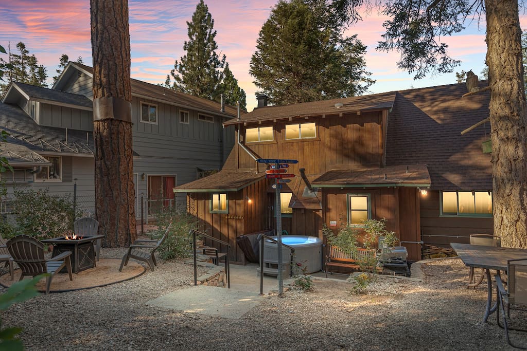 The outdoor space features a jacuzzi hot tub, surrounded by a gravel area with seating for relaxation. A propane fire pit is set nearby, with wooden benches situated around it. The cabin's multiple windows reflect the evening sky, contributing to a cozy and inviting atmosphere.