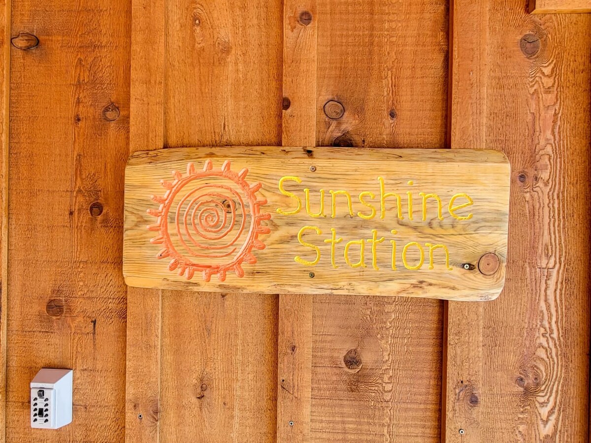 A wooden sign displaying the name 'Sunshine Station' is mounted on a rustic wooden wall. The sign features a sun design, with warm colors that complement the natural tones of the wood surrounding it.