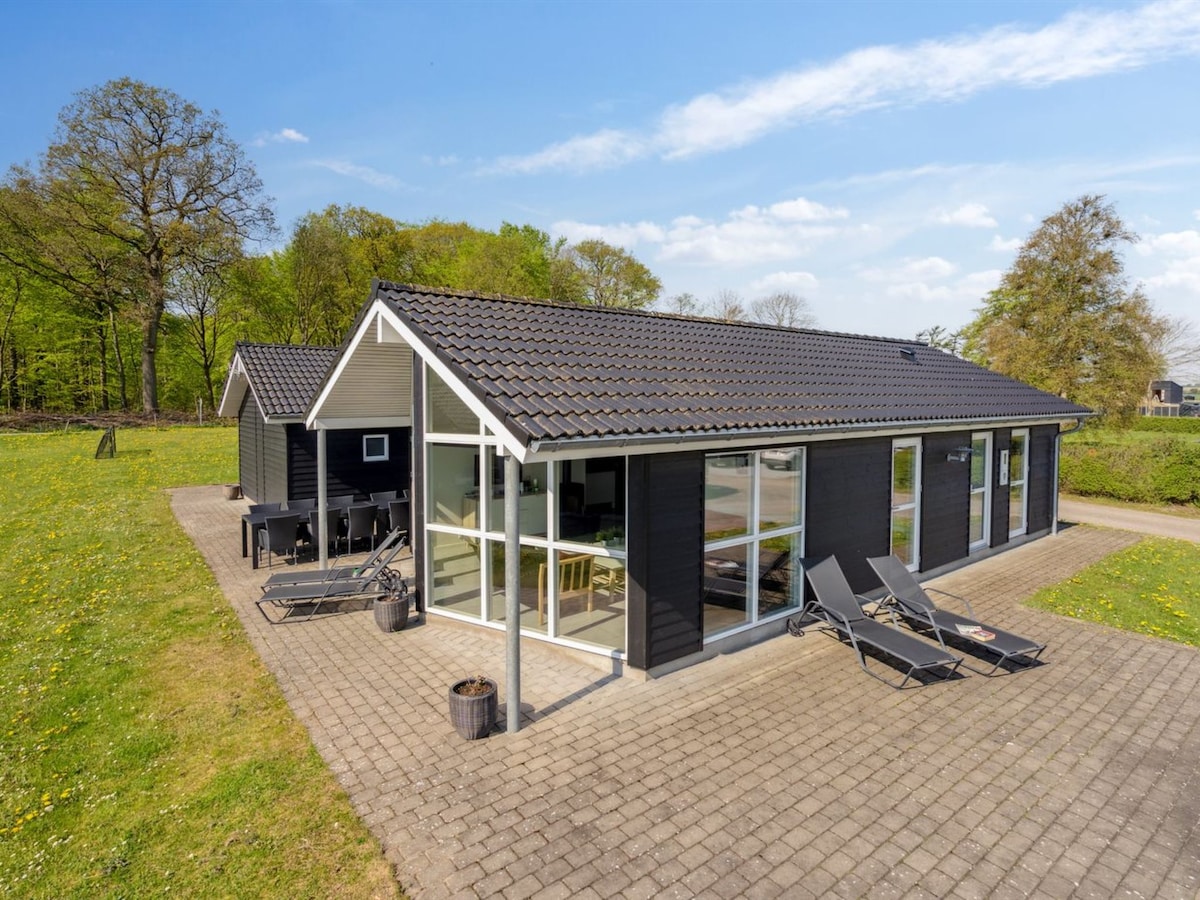 A modern single-family house is presented with a gabled roof and large windows. The outdoor space features a paved patio area with lounge chairs, surrounded by a grassy landscape and trees, creating a serene environment for relaxation.