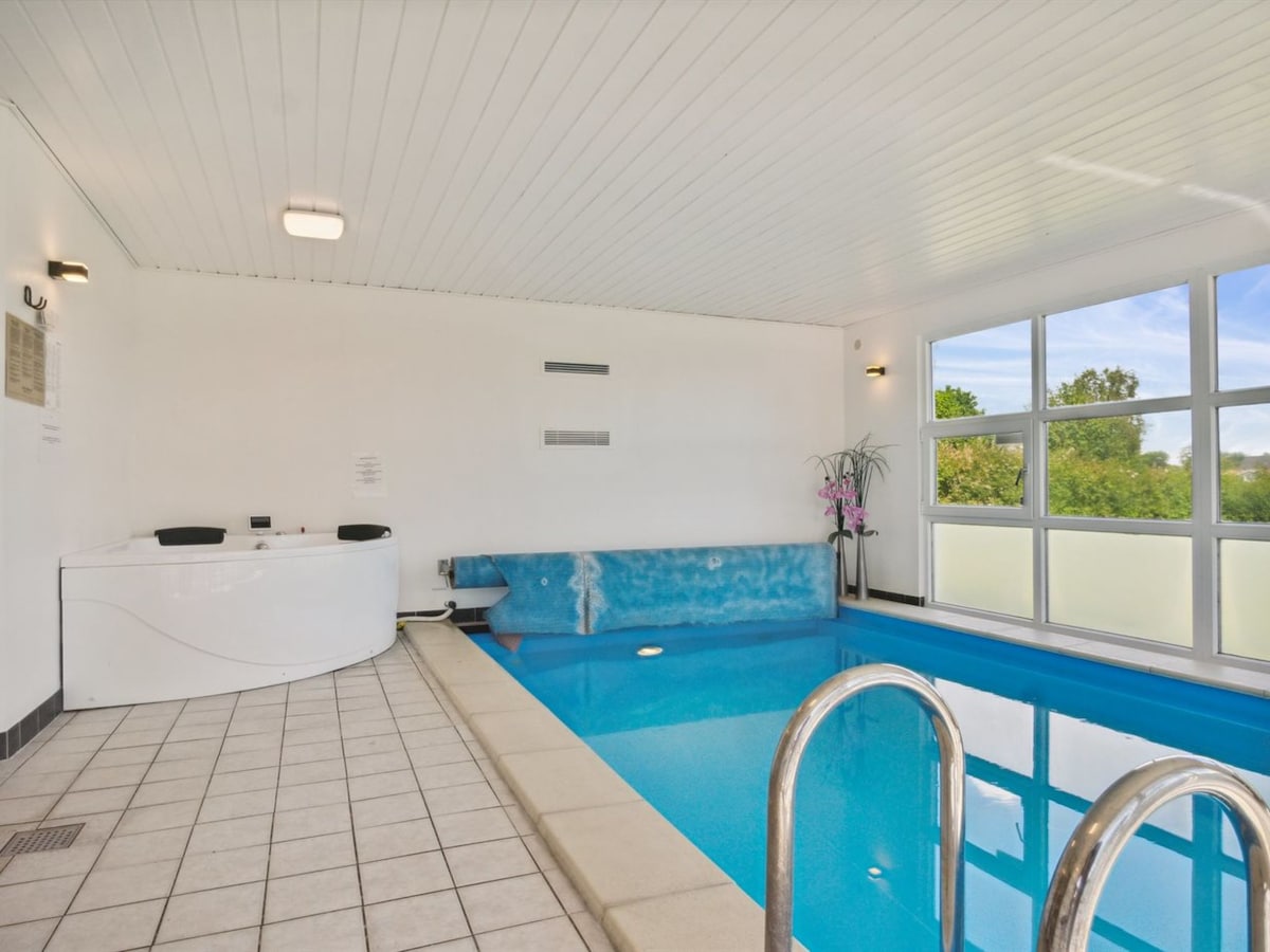 An indoor pool area is shown, featuring a rectangular pool with clear blue water. A white jacuzzi is positioned nearby, and large windows provide natural light and views of greenery outside. The floor is tiled in a light color, and a plant is placed in one corner.