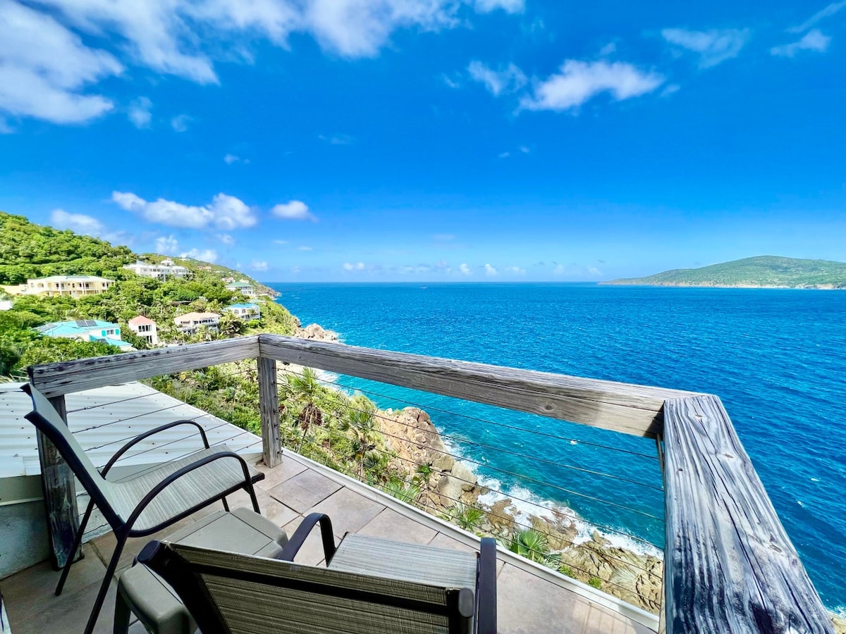 A wooden railing surrounds a patio area with two chairs, overlooking the sparkling blue ocean. The view extends to the distant green hills and the coastline, framed by a clear blue sky with scattered clouds.