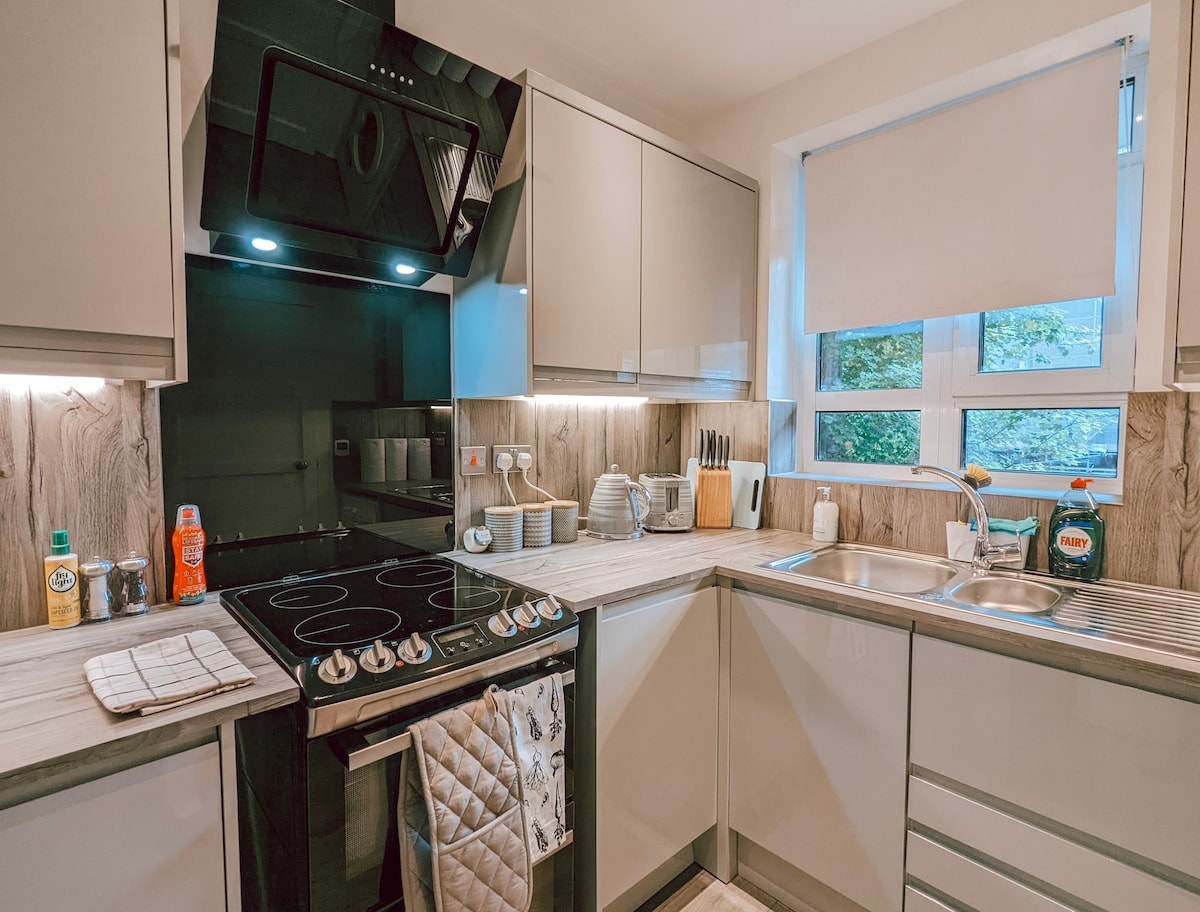 The kitchen features modern cabinetry with a sleek design, equipped with a gas stove, oven, and double sink. Countertops are lined with utensils, dishware, and cleaning supplies, while a window allows natural light to brighten the space.