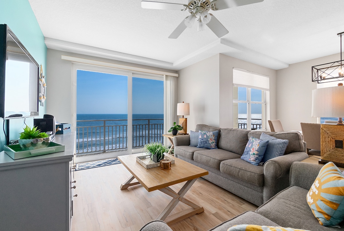 A living room area showcases comfortable seating arranged around a wooden coffee table. Expansive windows allow for a panoramic view of the ocean, with natural light illuminating the space. Decorative accents include cushions and a small plant atop a side table.