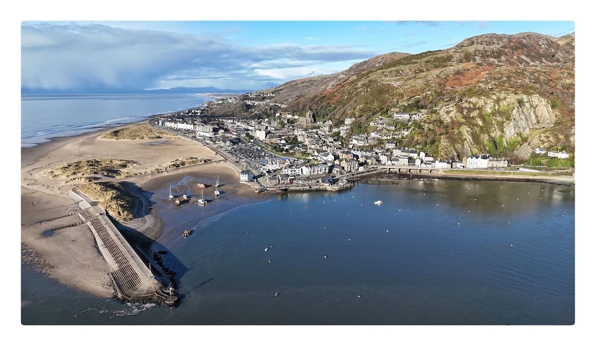 A panoramic view captures the coastal town nestled at the water's edge. Sandy beaches extend to the shoreline, while a series of buildings can be seen, accompanied by boats in the water. Hills rise in the background, creating a scenic landscape against a clear sky.