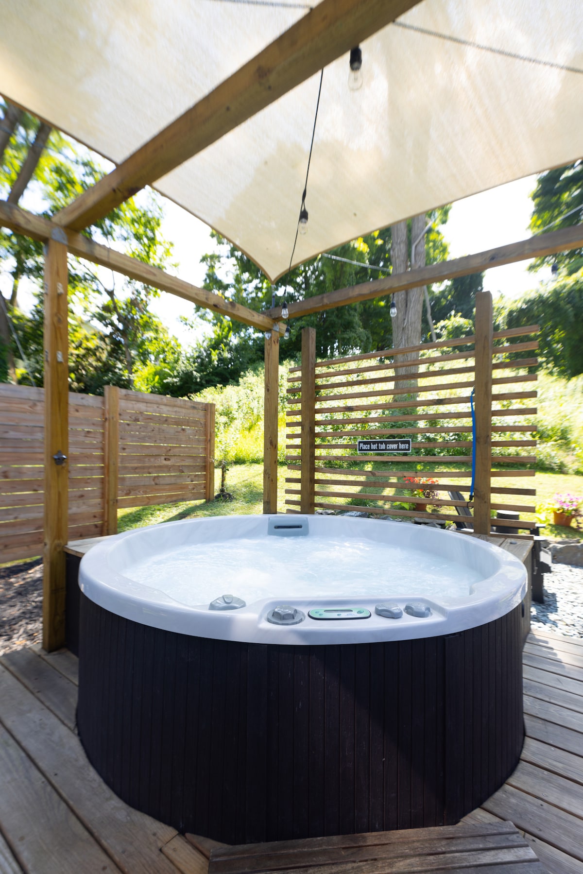 A private hot tub is surrounded by wooden decking and a shaded canopy, providing a tranquil spot to relax. Wooden slats frame the area, offering privacy while allowing natural light to filter through, complemented by lush greenery in the background.