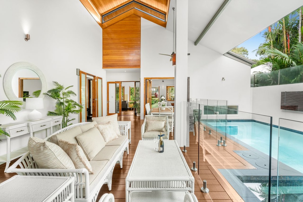 An open living area features high ceilings and a neutral color palette. Comfortable seating is arranged near large sliding doors that provide views of the lush landscaping and private internal pool. Natural light floods the space, creating a bright and airy atmosphere.