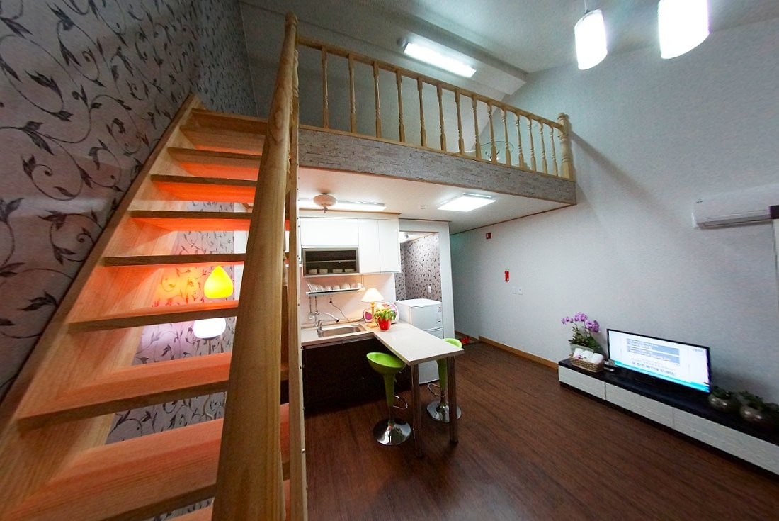 A two-level living area is showcased with wooden stairs leading to a loft space. The kitchen features modern appliances and a small dining table with green stools. Natural light fills the space, while decorative elements provide a cozy ambiance.