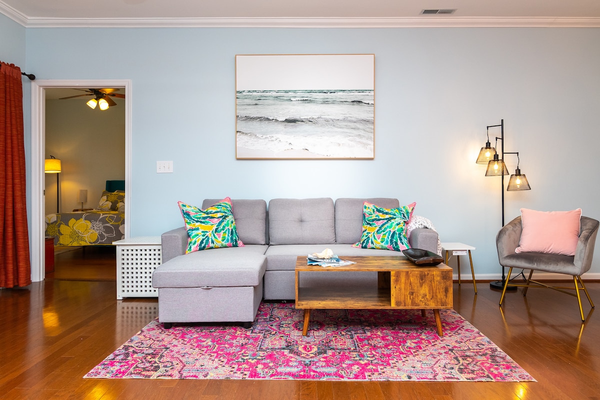 A spacious living area features a light grey sectional sofa adorned with colorful throw pillows. A wooden coffee table sits atop a vibrant area rug. A large wall art piece depicting ocean waves is displayed above the sofa, complemented by stylish floor lamps in the corner. Warm wood flooring enhances the inviting atmosphere.