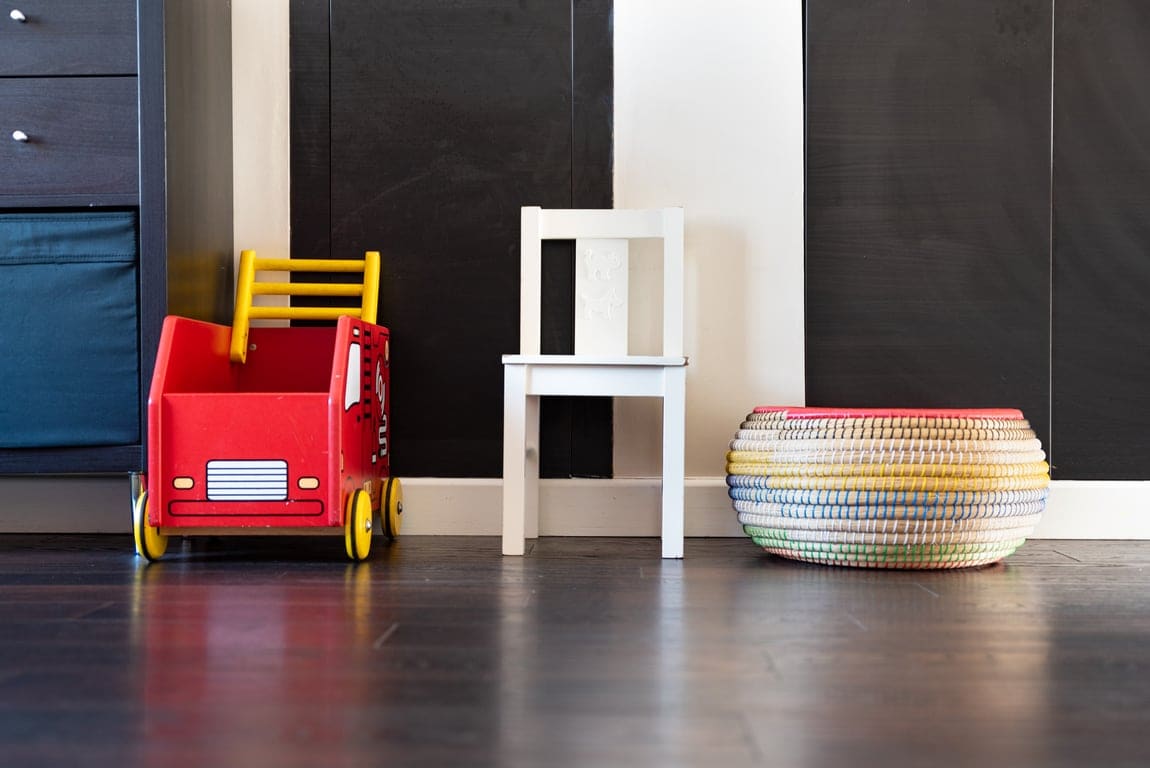 A vibrant red toy car sits beside a white chair, both positioned against a dark wall. A colorful woven basket is placed nearby, adding a playful touch to the room’s decor.