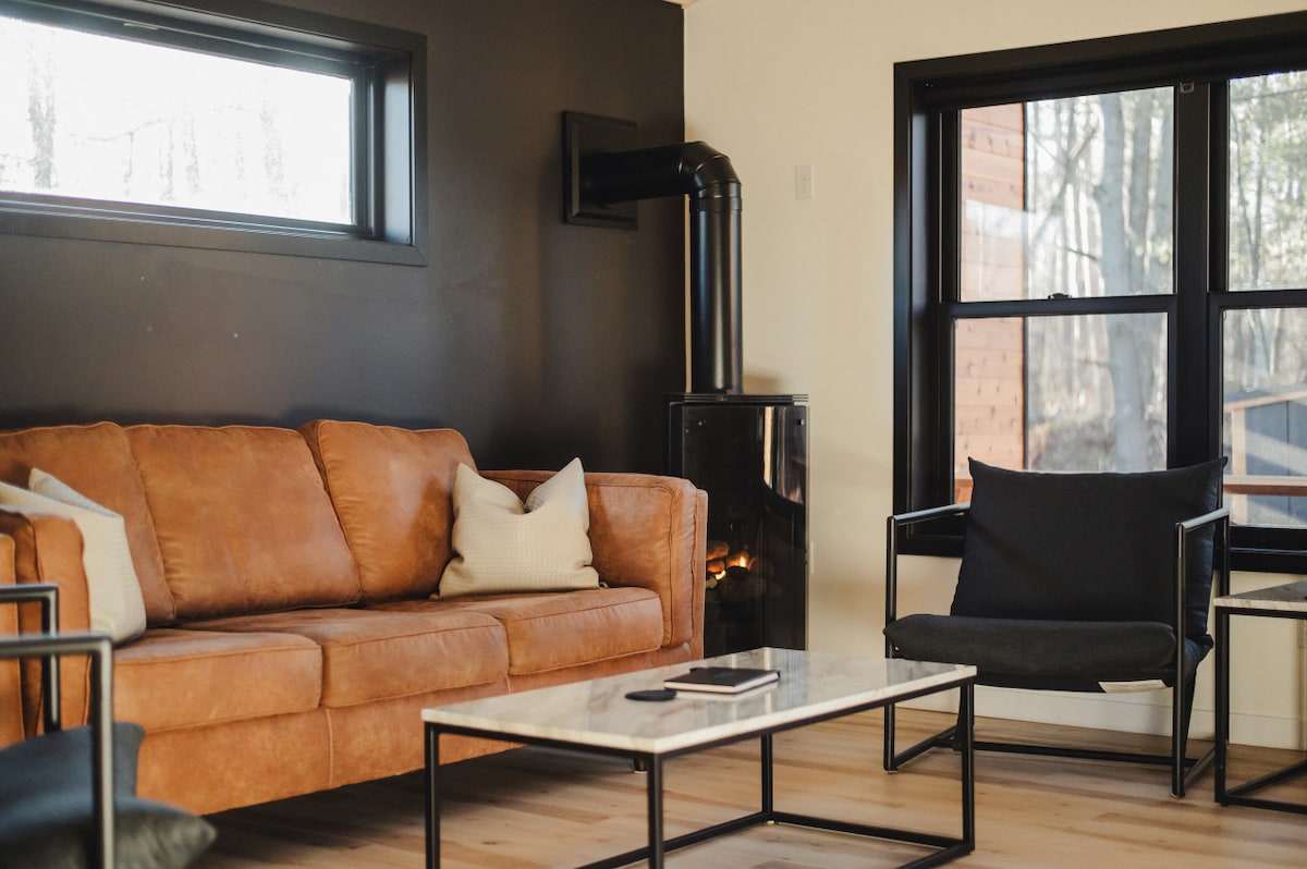 A modern living area features a brown leather sofa paired with two black armchairs. A rectangular coffee table sits in the center, while a sleek black stove is visible in the corner. Large windows allow natural light to fill the room, showcasing outdoor scenery.