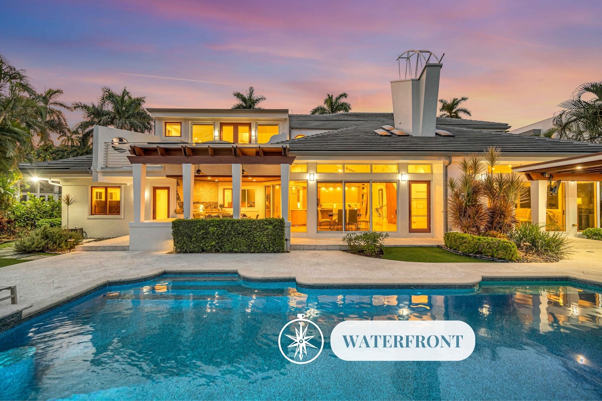 The exterior of the villa is showcased, featuring a sleek design that opens to a heated pool area. Lush landscaping surrounds the water's edge, providing a tranquil atmosphere. The setting is enhanced by a beautiful sunset sky, adding to the allure of this waterfront property.