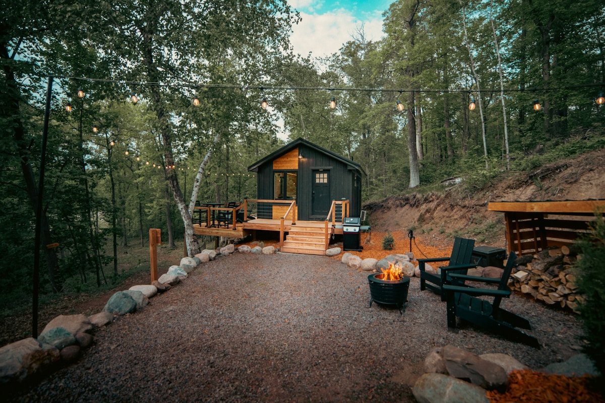 A charming tiny house is nestled among the trees, featuring a welcoming deck with steps leading up to the entrance. A gravel path is lined with stones, and a fire pit accompanied by seating is positioned nearby, creating a cozy outdoor gathering space.