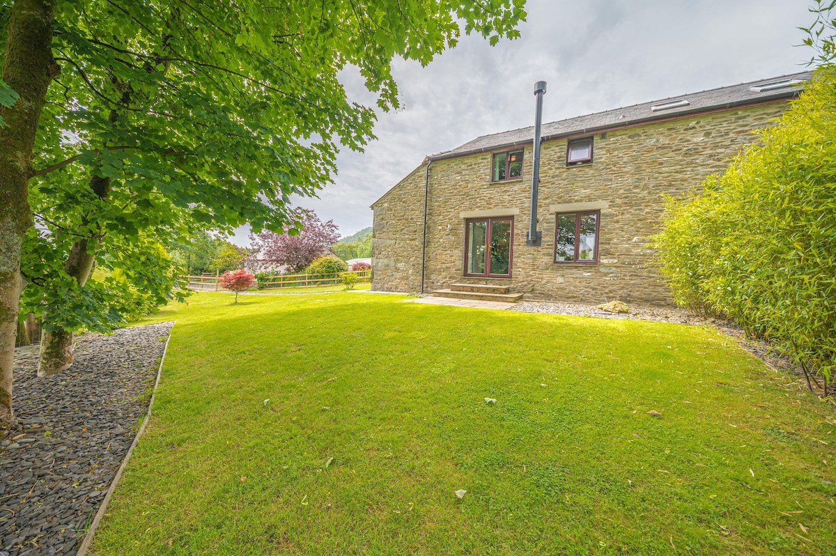 A stone cottage is positioned amidst a green lawn, framed by leafy trees. The exterior features large windows, and a central door leads onto a small patio area. The peaceful garden is landscaped with gravel pathways and a variety of plants, offering a scenic view of the countryside.