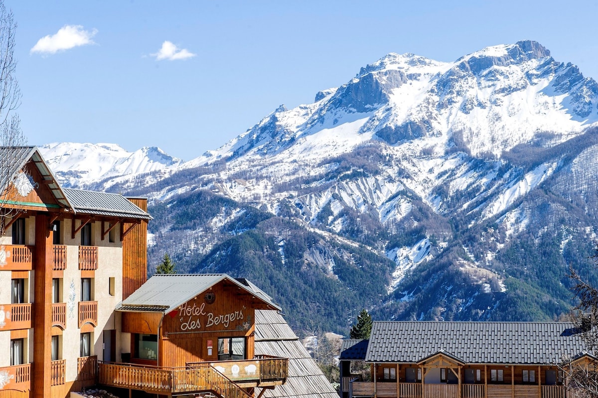 The image showcases a charming alpine hotel with wooden balconies and a sloping roof, set against the backdrop of snow-capped mountains and a clear blue sky. The surrounding landscape features evergreen trees, enhancing the serene mountain atmosphere.
