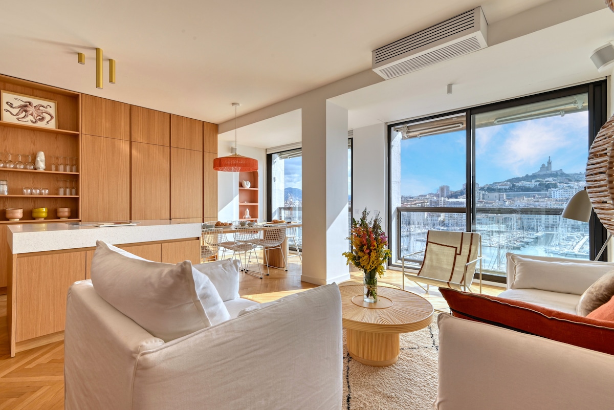 A spacious living area features large bay windows with views of the port and city. Natural light highlights the modern furniture, including a round coffee table and comfortable seating. The open kitchen, adorned with wooden cabinetry, complements the inviting atmosphere.