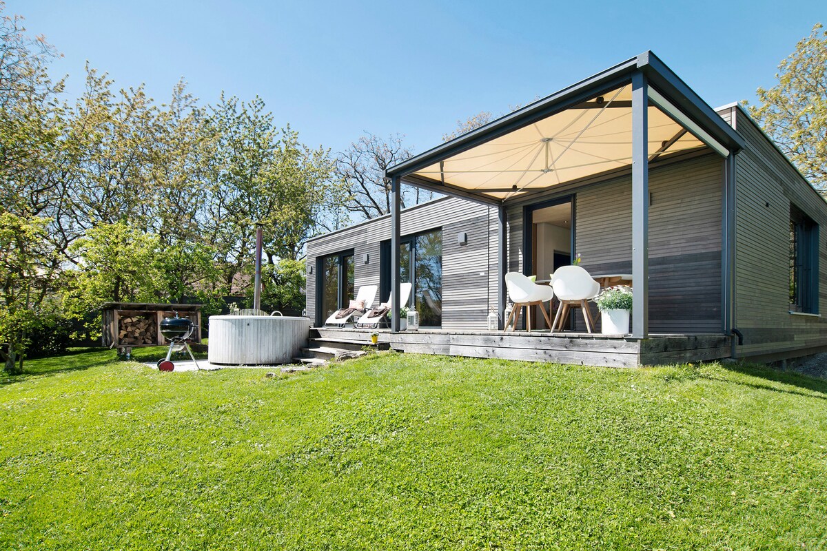 The exterior of the chalet presents a spacious deck with modern seating under a large canopy. A private jacuzzi is positioned nearby amidst the well-maintained lawn, surrounded by greenery. Large glass doors allow for easy access to the interior.