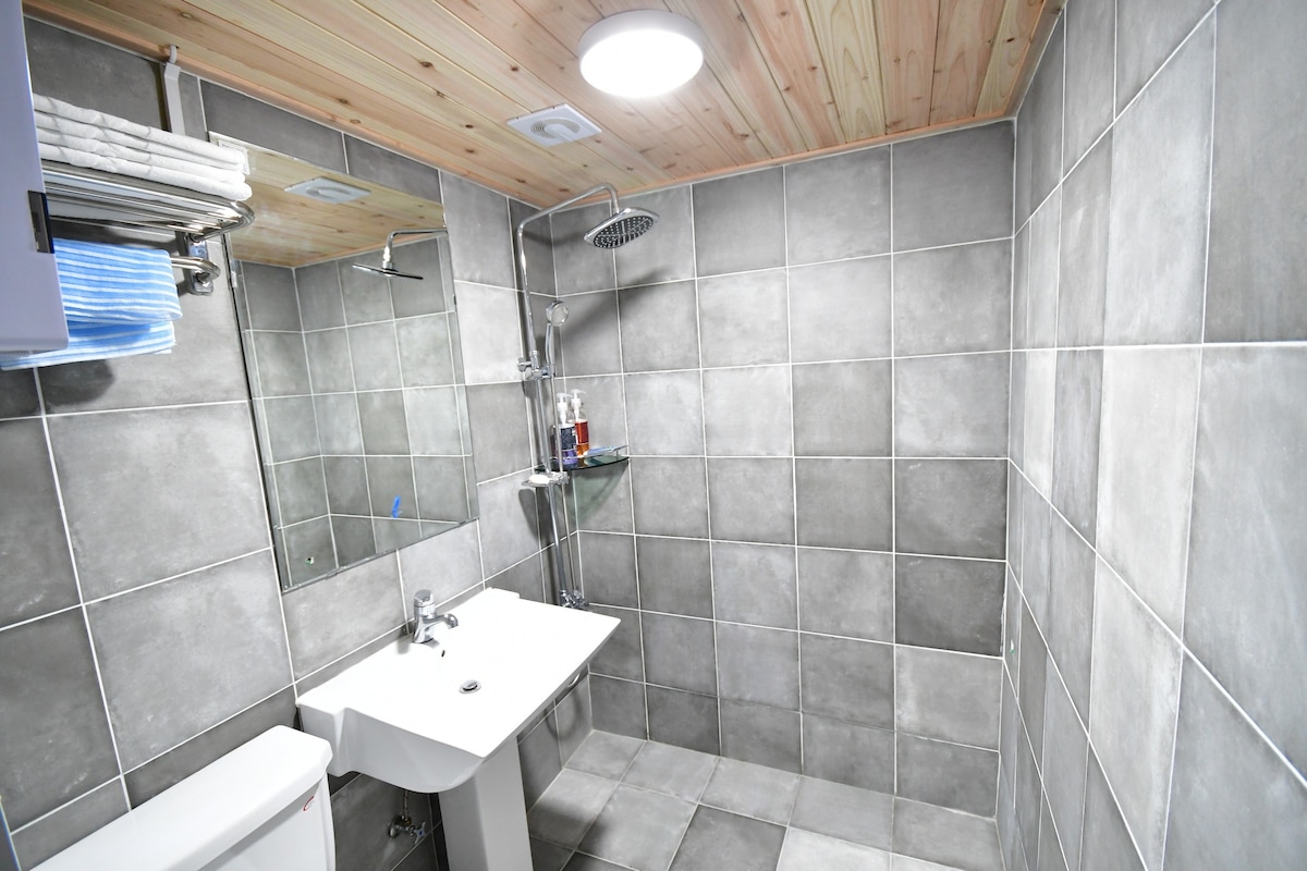The bathroom features modern gray tiled walls and flooring. A glass shower stall is equipped with a detachable showerhead. A rectangular sink sits beneath a wall-mounted mirror, with a shelf above displaying neatly arranged towels.