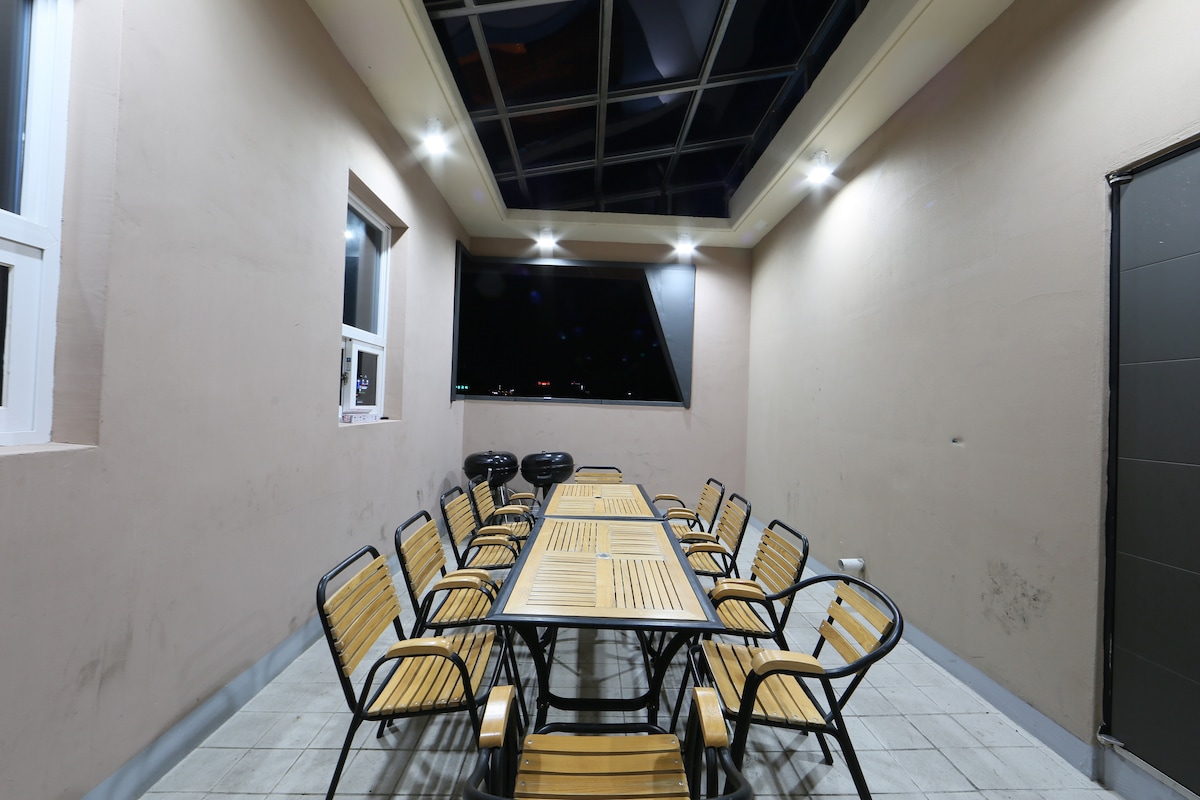 An outdoor dining area features a long wooden table surrounded by multiple wooden chairs. The space is illuminated with overhead lights and has a modern glass ceiling, creating a bright atmosphere for gatherings.