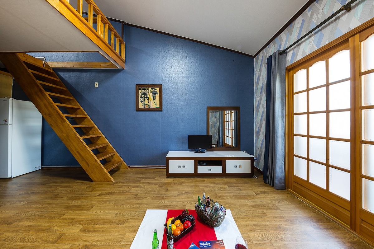 An open living area showcases wooden flooring and blue accent walls. A staircase leads to the upper level, while a low white furniture piece supports a television. A mirror reflects the space, complemented by a red tray adorned with a variety of beverages and snacks.