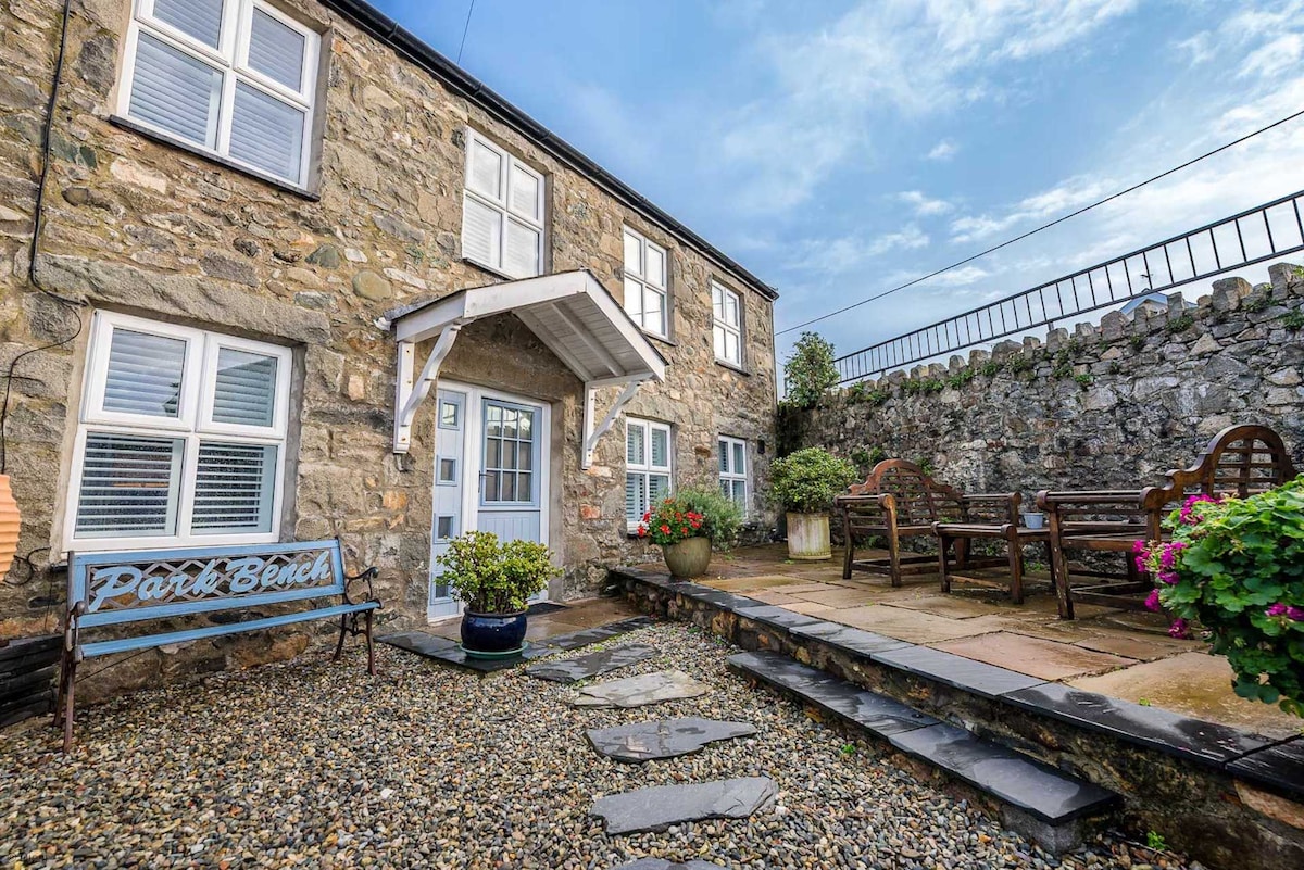 The entrance of the cottage is shown, featuring a pale blue door under a covered porch. The front garden includes decorative gravel, stepping stones, and a stone wall. A comfortable seating area is arranged with wooden furniture, surrounded by potted flowers.