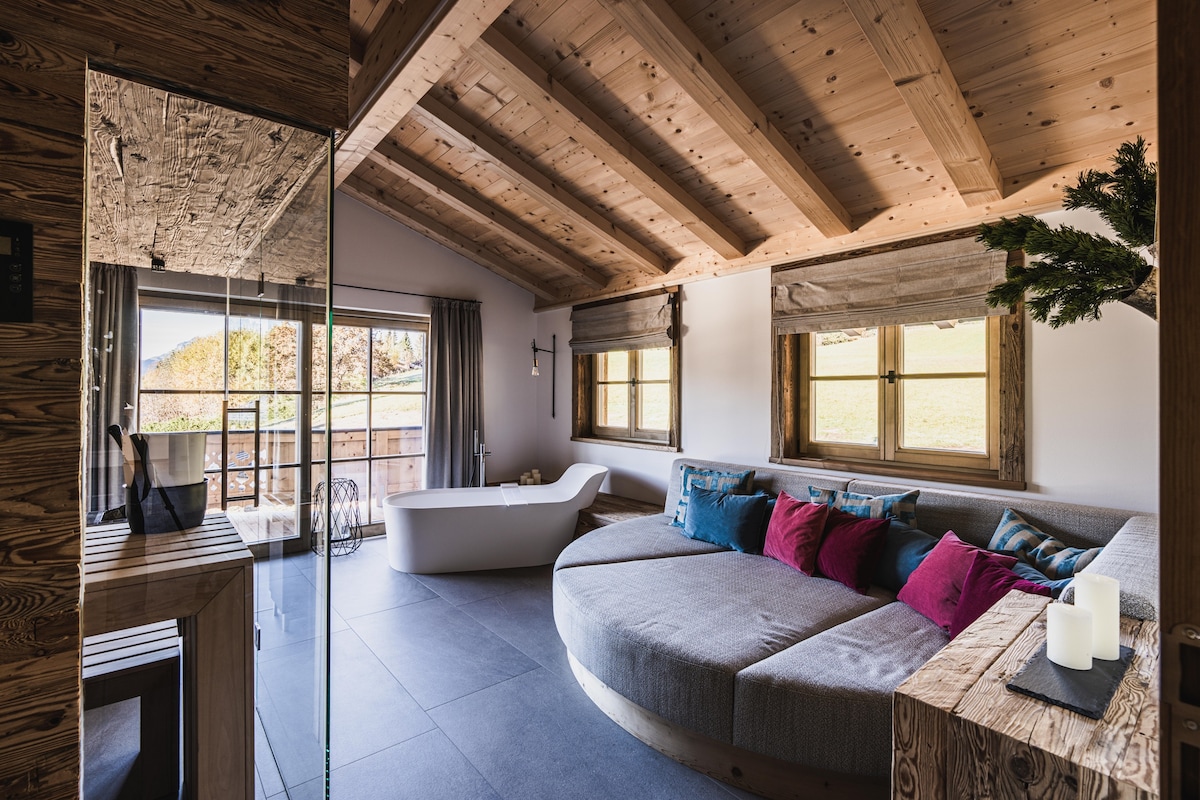 A spacious living area is highlighted by a unique circular sofa adorned with colorful pillows. Natural light filters through large windows, offering views of the outdoors. The wooden ceiling beams complement the contemporary design, with a freestanding bathtub adding a touch of luxury.