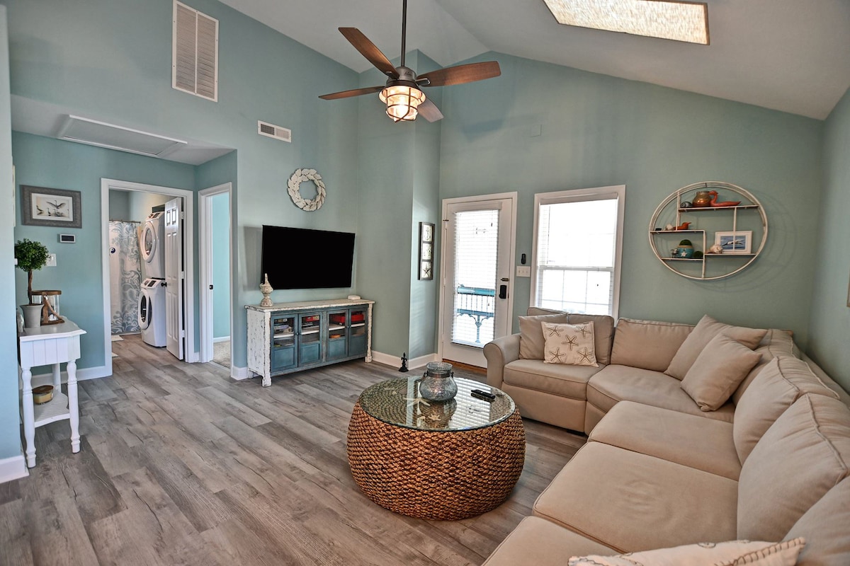 The living area features a spacious sectional sofa arranged around a round wicker coffee table. A large smart TV is mounted on the wall, opposite the seating. Light blue walls and wood-like flooring create a calming ambiance, complemented by natural light through the nearby windows.