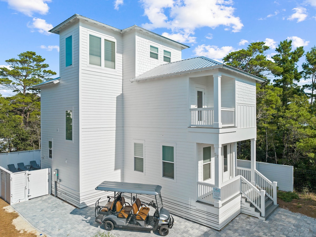 The exterior of a newly built coastal house is shown, featuring a light-colored facade with multiple windows allowing natural light. A golf cart is parked on a paved area, providing easy access to nearby attractions. Surrounding trees enhance the private setting.