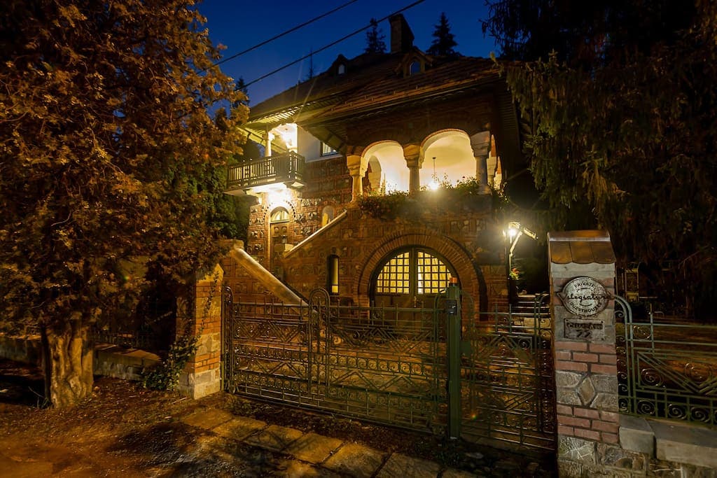 The villa's exterior is elegantly illuminated at night, highlighting its Neo-Romanian architectural style. Intricate wrought iron gates and stone pathways lead to the grand entrance, flanked by lush trees and soft lighting that accentuate the villa's charm.