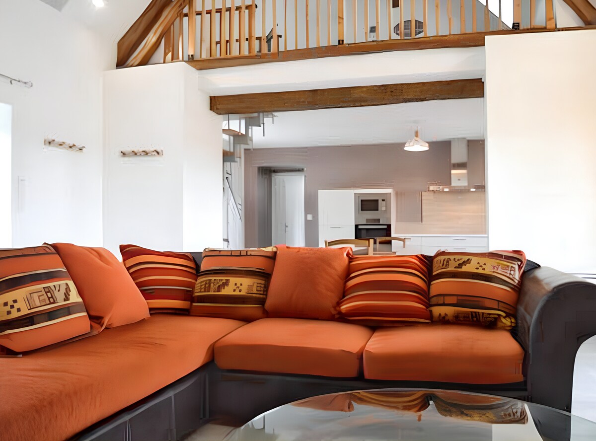 A spacious living area features a large orange sectional sofa adorned with patterned throw pillows. A coffee table occupies the center, and a glimpse of an open kitchen and dining area is visible in the background, showcasing a modern and inviting design.