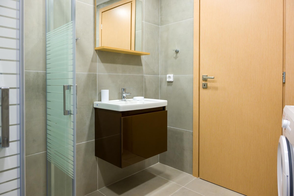 A modern bathroom is featured with neutral grey tiles. A sleek, dark vanity unit houses a white sink, complemented by a large mirror above. A glass shower stall stands in one corner, and a separate wooden door leads to the hallway.