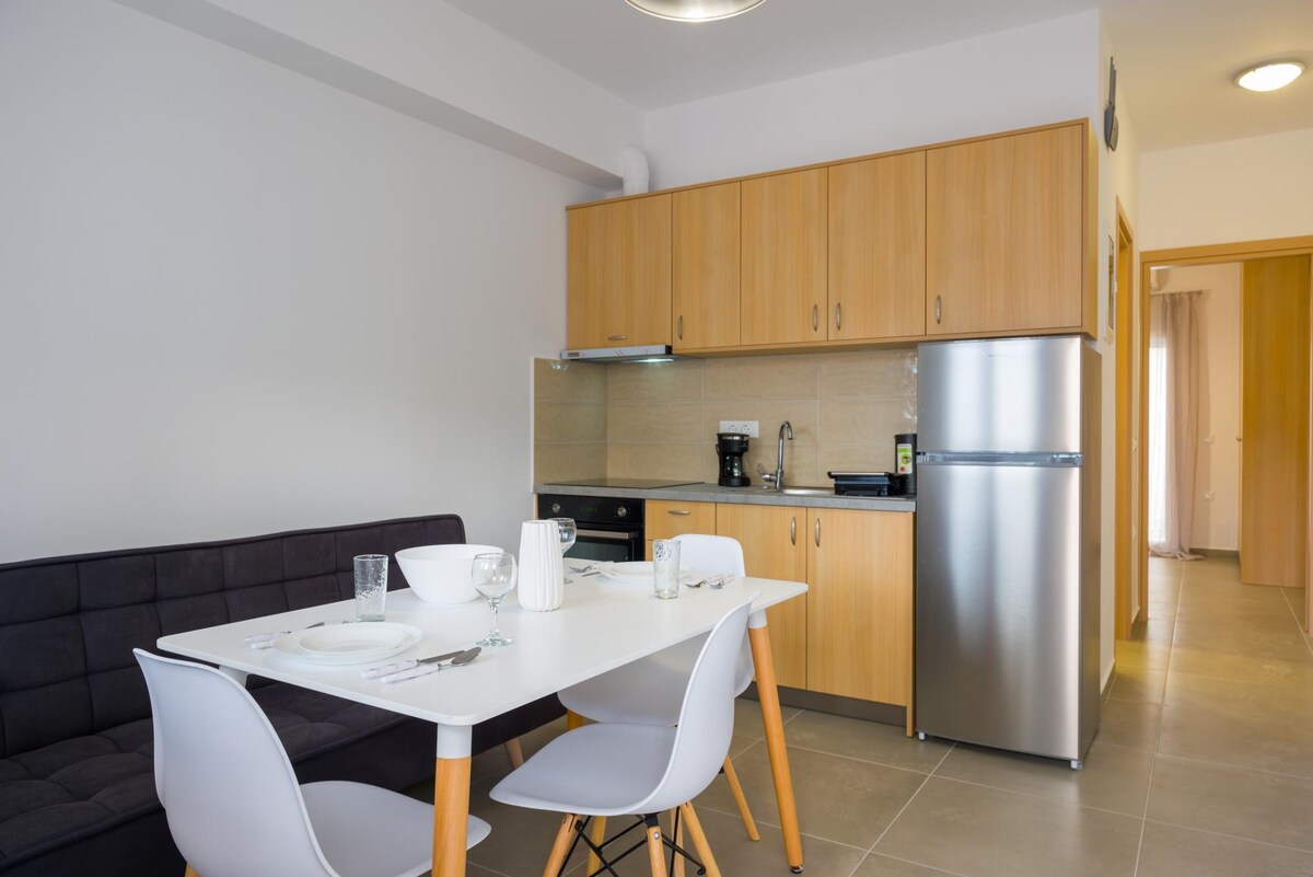 A dining area features a white table set for four, accompanied by modern chairs. A kitchenette with wooden cabinetry and stainless steel appliances is visible in the background, including a refrigerator and coffee maker. Natural light illuminates the space through nearby windows.
