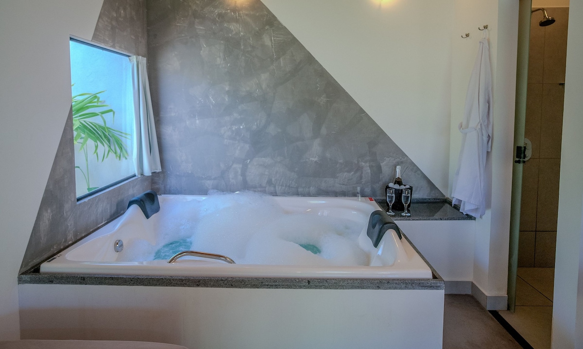 A spacious jacuzzi bathtub for two is positioned next to a window framed by light curtains, allowing natural light to enhance the tranquil atmosphere. The sleek, modern design features stone accents and is complemented by a drink set on the countertop nearby.