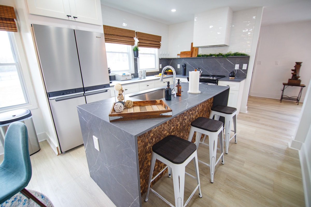 A modern kitchen features a large island with a stone countertop, complemented by three wooden stools. Stainless steel appliances and white cabinetry are included, with natural light flowing through windows adorned with wooden blinds. Decorative elements are arranged on the island, enhancing the space.