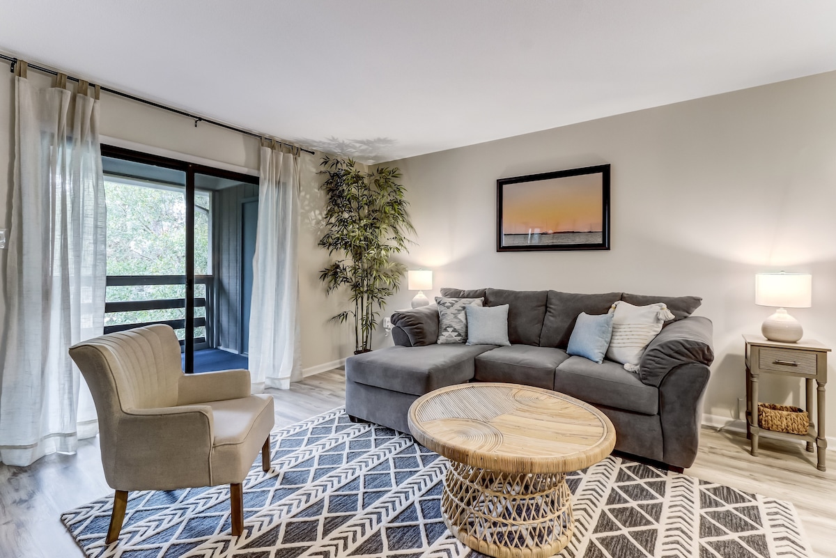 A spacious living room features a comfortable sofa with an attached chaise lounge, complemented by a stylish chair. Natural light filters through sheer curtains, illuminating the decor. A round coffee table sits centrally, and a sliding glass door leads to the screened balcony, enhancing the connection to the outdoors.