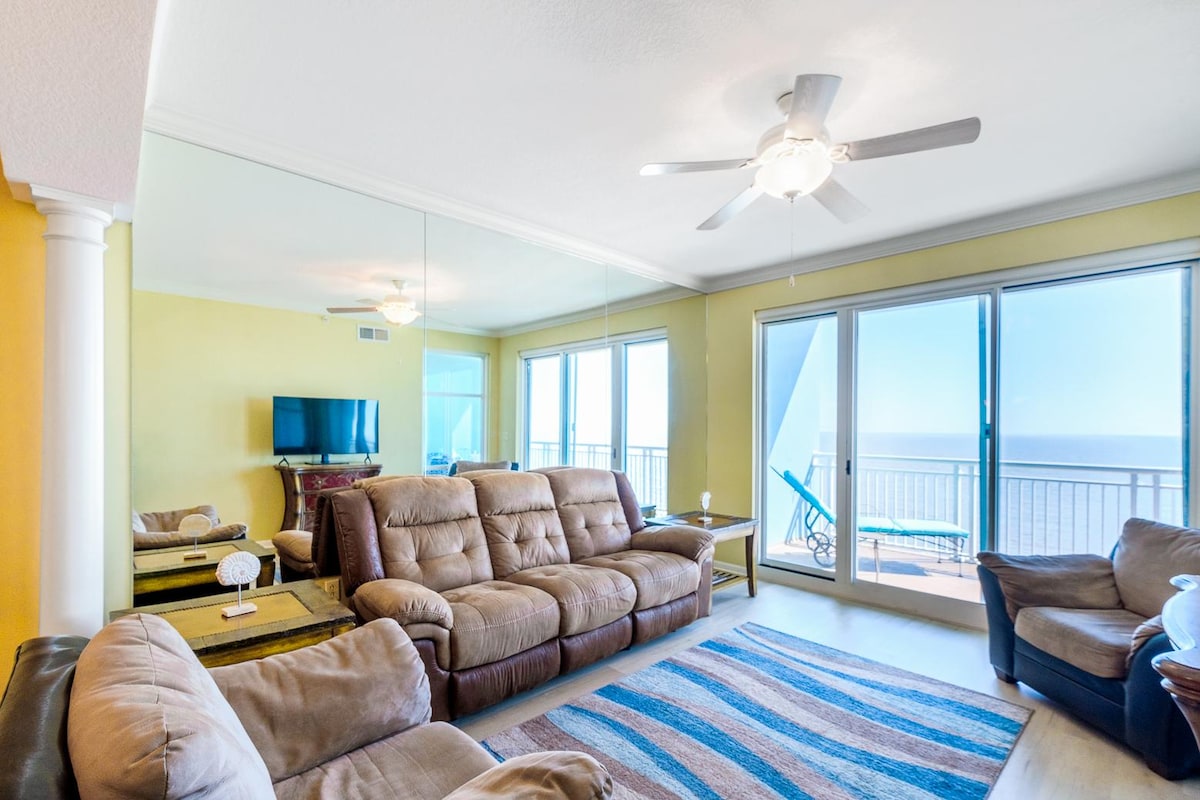 A spacious living area features comfortable seating with a large sectional sofa and armchair. Light colors brighten the space, complemented by a ceiling fan. Expansive glass doors provide access to a private balcony with views of the water, enhancing the inviting atmosphere.