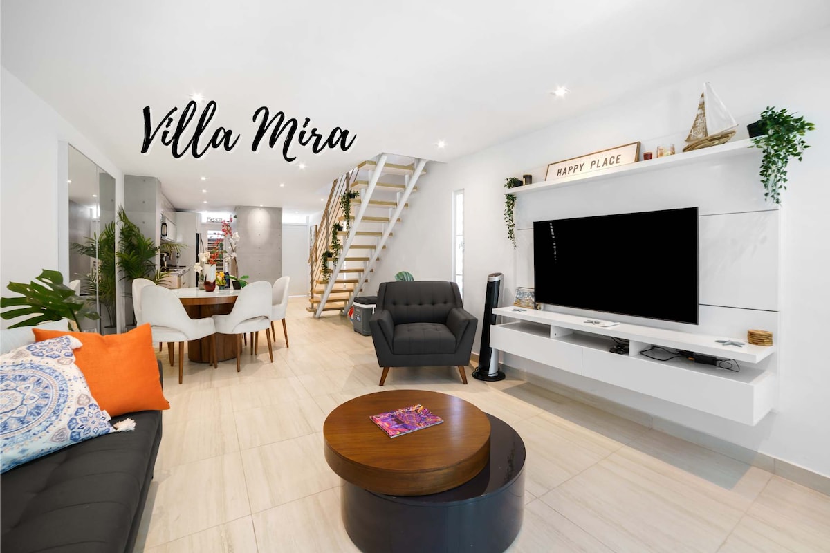 A modern living space features a mix of comfortable furniture, including a deep-cushion couch and an armchair, with a round coffee table in the center. A large flat-screen TV is mounted on a white shelf, and a dining area is visible in the background.