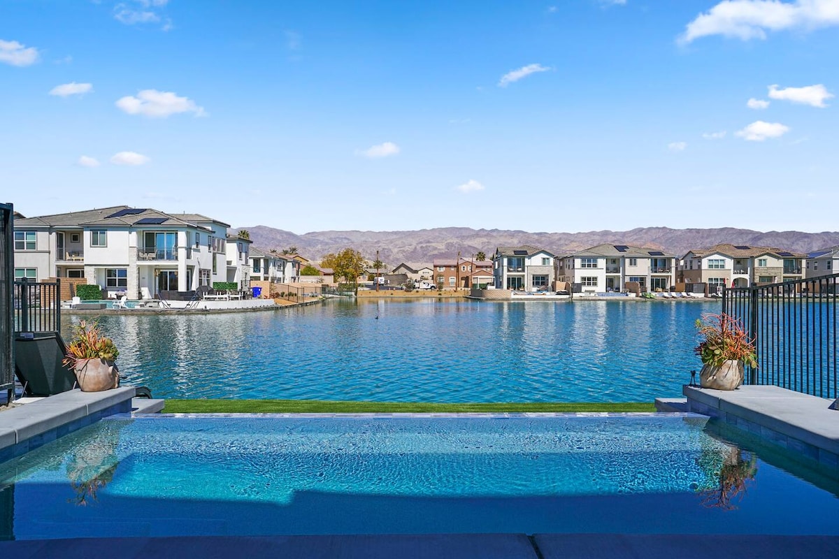 A serene view of a sparkling infinity pool overlooks a tranquil lake, framed by elegant landscaping. The clear blue sky extends above, with distant mountains creating a picturesque backdrop. Nearby homes are visible, reflecting a comfortable community atmosphere.
