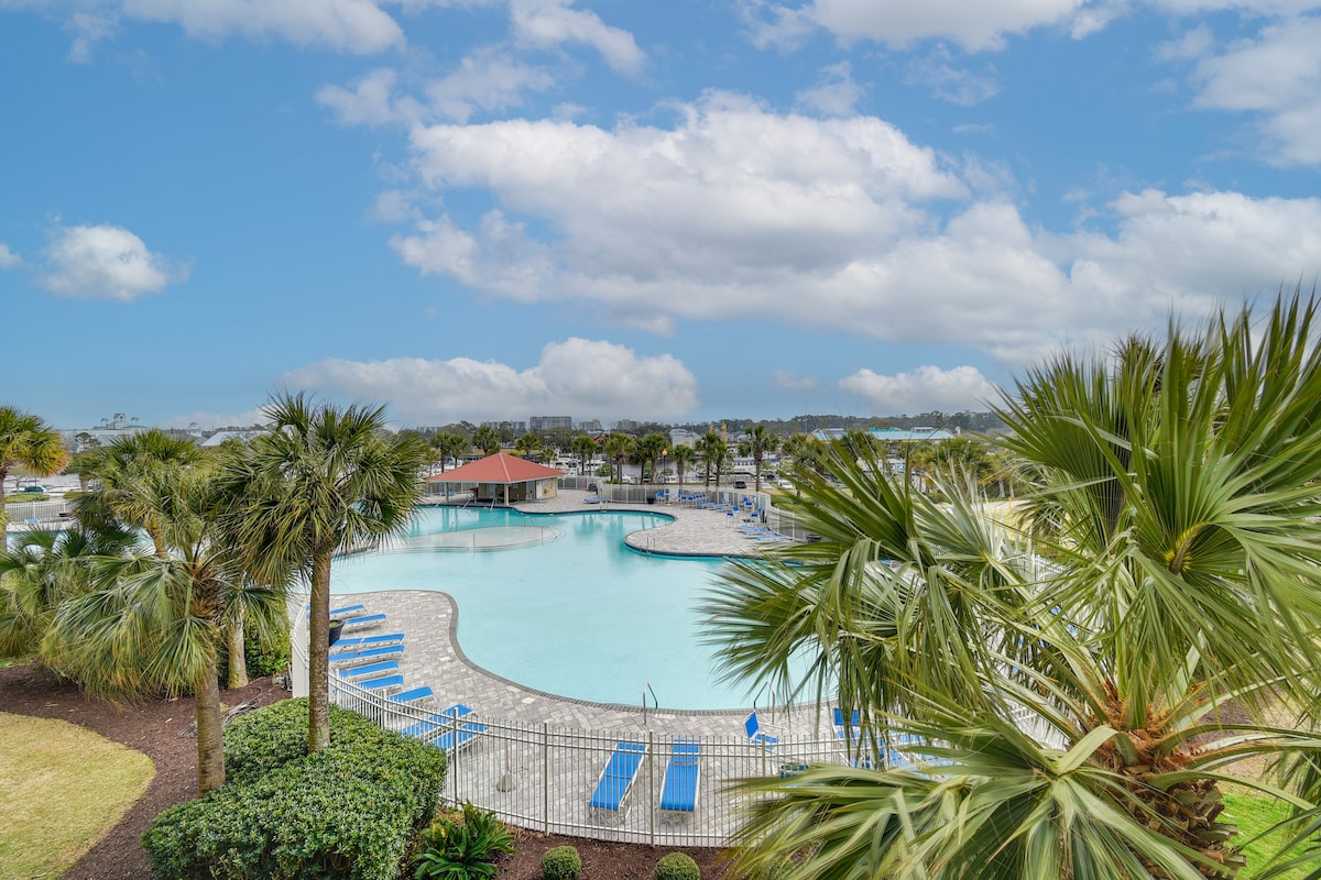 A spacious community pool is surrounded by manicured landscaping, featuring palm trees and lounge chairs. The water’s surface reflects the clouds above, while a covered cabana is visible in the background, providing a shaded area for relaxation.