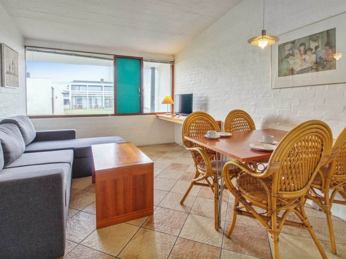 A bright living area is present, featuring a comfortable gray sofa and a wooden coffee table. A dining table for four is surrounded by wicker chairs. A window allows natural light to fill the space, with a TV positioned nearby.