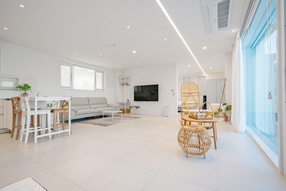 A bright and airy living space features a comfortable sofa and a flat-screen television mounted on the wall. A dining set for four is positioned near the window, enhancing the openness of the room. Natural light is welcomed through large windows.